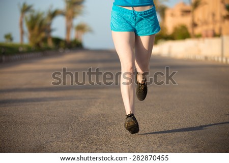 Healthy lifestyle: close up of young woman legs in running shoes jogging on fast speed outdoors on the sunny summer street in tropics, palm trees on the background