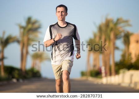 Healthy lifestyle: positive sporty young man working out outdoors in the summer sunny street in tropics, jogging on the road, palm trees on the background