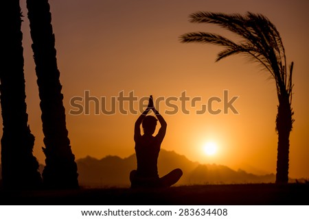 Silhouette of young woman sitting in serene picturesque place among palm trees, watching sun rising or setting in mountains, meditating in sukhasana easy pose