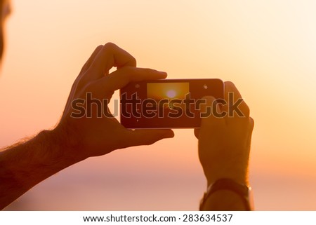 Male hands holding mobile phone with image of sunset or sunrise on the screen, taking photo in picturesque sea scenery, back view, close up