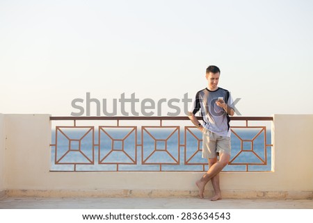 Minimalistic portrait of young happy smiling man holding cellphone, using app, making call, messaging text or dialing number, standing barefoot on summer street with sea scenery, copy space