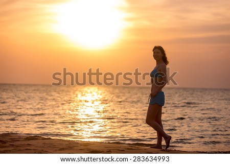 Smiling young woman walking on the sand beach at picturesque colorful sunrise or sunset, lit by warm sunlight, copy space