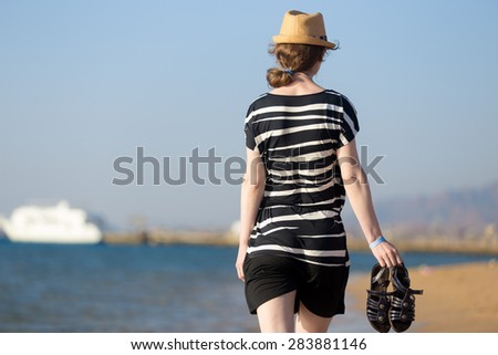 Young woman in short dress fluttering in sea breeze walking at sea coast holding her blue leather sandals, back view