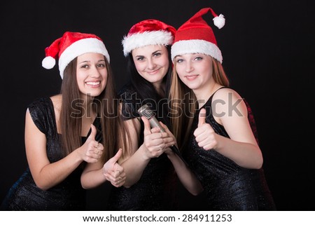 Group of three positive, happy smiling beautiful girls vocalists posing in cute red santa claus hats with microphone, showing \