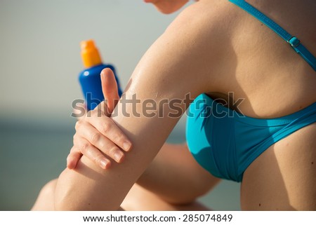 Young woman in blue swimwear on the beach holding bottle of suntan lotion, applying sun cream on her arm before tanning, close up