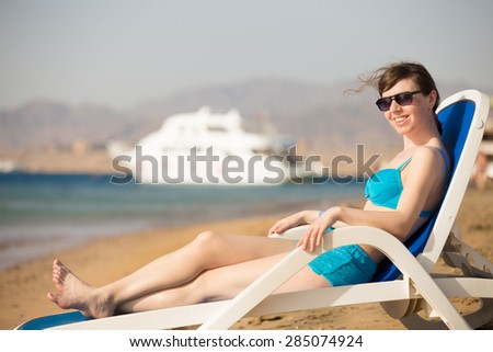 Young slim happy smiling woman in blue swimwear and sunglasses relaxing on sun lounger at sand beach, tanning in sunlight on tropical sea bay, white yacht on the background
