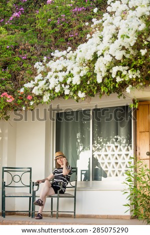 Young woman in cute straw hat and summer dress sitting on chair on terrace of white cottage house with flower roof, using cell phone, talking on phone, making call