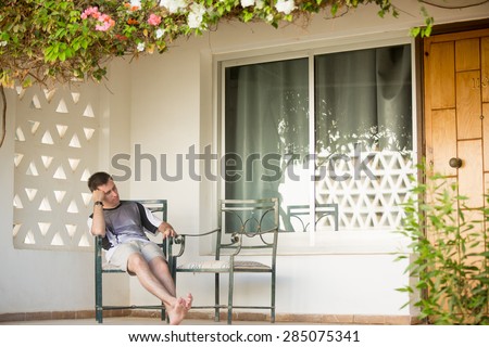 Young handsome man sleeping on chair on the porch of white cottage house covered in flowers