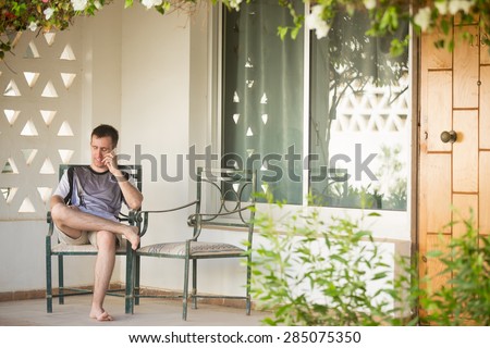 Young handsome smiling man sitting barefoot in relaxed pose on chair on the porch of white summer cottage house, holding cell phone, making call, talking on smartphone