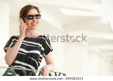 Portrait of young happy smiling woman in sunglasses and black and white summer dress standing in a building with white stucco walls, talking on cellphone, making call, copy space