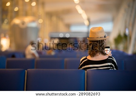 Young traveling woman waiting for transYoung female traveler in summer dress and straw hat waiting for trip, sitting with smartphone in modern station lounge area with blue seats, back viewport