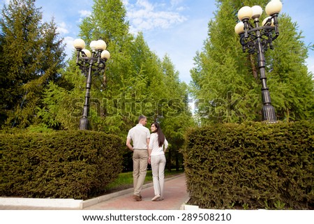 Couple of lovers, attractive young man and woman on date, walking in park with trimmed bushes, holding hands, smiling girlfriend looking back, full length, rear view