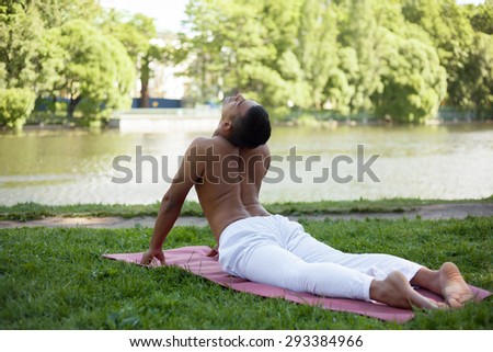 Indian young man in white linen clothes practicing yoga, fitness, pilates on riverbank in park, lying in bhudjangasana (Cobra Pose), full length, back view