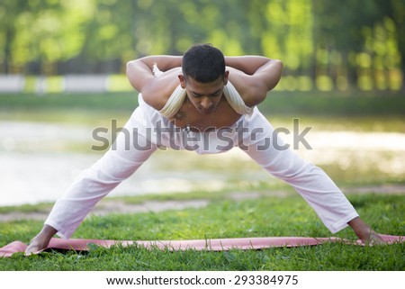 Attractive Indian young man in white linen clothes working out on riverbank in park, doing Wide Legged Forward Bend, Ardha Prasarita Padottanasana variation, hands behind back, full length