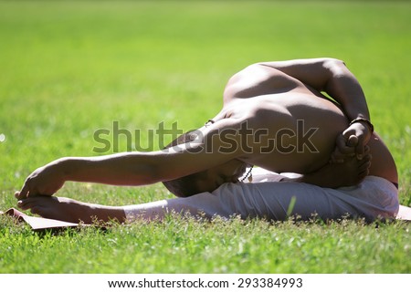 Serene Indian young man in white pants practicing yoga, fitness or pilates on green grass in park, doing ardha baddha padma paschimottanasana, half bound lotus forward bend, full length