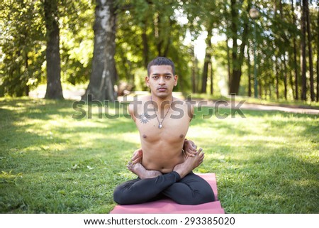 Serene sporty Indian young man working out on red mat on forest lawn, sitting with crossed legs, doing Baddha Padmasana, Bound or Locked Lotus pose, full length, front view