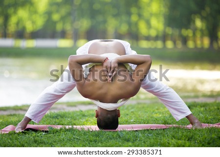 Attractive Indian young man in white linen clothes practicing yoga, fitness, pilates on lake in park, doing Wide Legged Forward Bend, Prasarita Padottanasana variation, hands behind back, full length