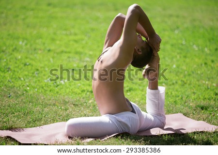 Serene attractive Indian young man in white pants doing stretching sport exercises on green lawn outdoor, practicing yoga, fitness or pilates, sitting in One Legged King Pigeon Pose 1