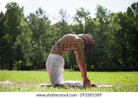 Serene Indian young man in white pants doing stretching sport yoga, fitness or pilates exercises on green grass, working out for flexible spine, stands on knees in ushtrasana, Camel Pose