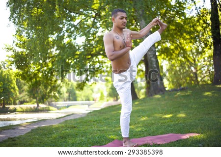 Serene attractive Indian young man in white linen clothes working out on river bank in park, standing in Extended Hand to Big Toe yoga pose, Utthita Hasta Padangushthasana, full length