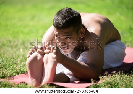 Handsome Indian young yogi man in white pants practicing yoga, fitness or pilates on red mat on grass in park, doing stretching exercises, seated Forward Bend pose, Paschimottanasana, full length