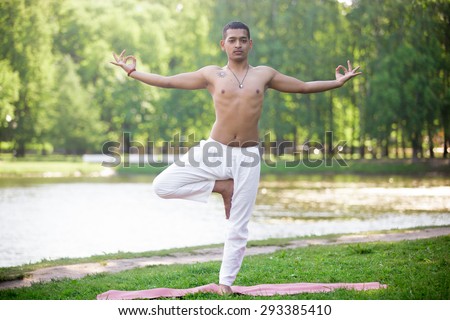 Serene attractive Indian young man in white linen clothes working out on river bank in park, standing in Vrikshasana (Vriksasana, Tree Pose) with hands in jnana mudra, full length