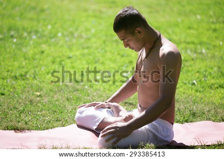 Serene Indian young man in white pants practicing yoga on green grass in park, doing Padmasana, sitting with closed eyes in Lotus Pose, breathing, meditating, relaxing, full length