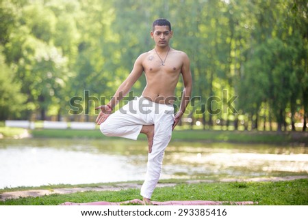 Serene attractive Indian young man in white linen clothes working out on lake in park, standing in Vrikshasana (Vriksasana, Tree Pose), full length