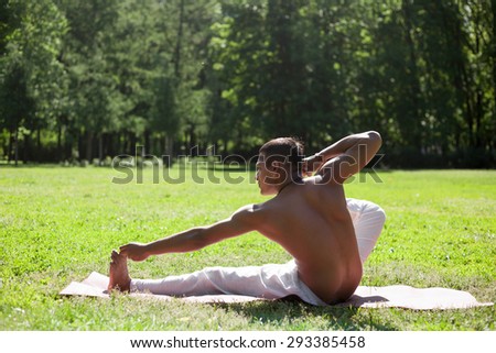 Profile of serene Indian young man in white pants practicing yoga, fitness or pilates on green grass in park, doing Akarna Dhanurasana I, Archer Pose