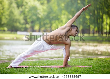 Profile of attractive Indian young man in white linen clothes practicing yoga, fitness, pilates on lake in park, standing in Utthita Parsvakonasana, Extended Side Angle Pose, full length