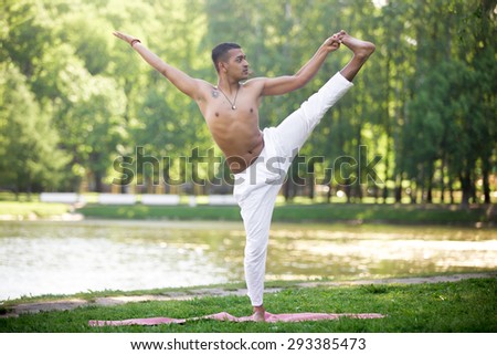 Attractive Indian young man in white linen clothes practicing yoga, fitness, pilates on riverbank in park, standing in Extended Hand to Big Toe yoga pose, Utthita Hasta Padangustasana, full length