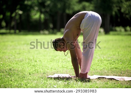 Profile of sporty young man exercising in park, doing yoga, fitness or pilates training, standing in uttanasana (forward fold, head to knees pose), surya namaskar sequence, sun salutation complex
