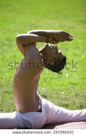 Portrait of handsome sporty Indian young man exercising on grass in park, doing yoga, fitness or pilates training, sitting in One Legged King Pigeon Pose with crossed arms, close up