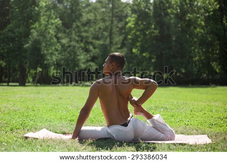 Handsome sporty Indian young man in white pants working out on grass in park, doing yoga, fitness or pilates training, sitting in One Legged King Pigeon Pose 1, back view