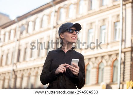 Young smiling traveler woman wearing casual clothes, cap and sunglasses holding cellphone, using app, gps, searching for direction while walking in European city street in summer