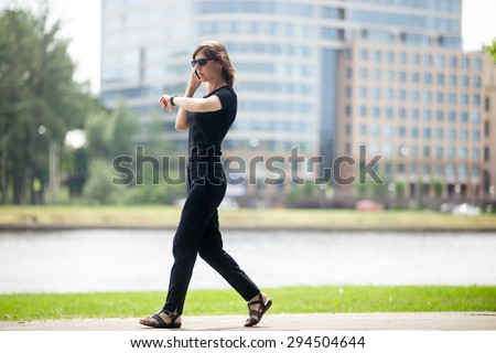 Young confident business woman walking at fast speed, looking at wristwatch, talking on cellphone on the city street in front of blue glass modern office building beside riverbank, full length