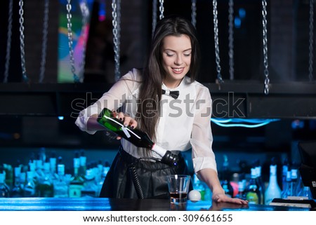 Beautiful cheerful brunette bartender girl in white shirt and black bow tie, serving alcohol drink at nightclub bar, holding bottle in hand, pouring drink in glass