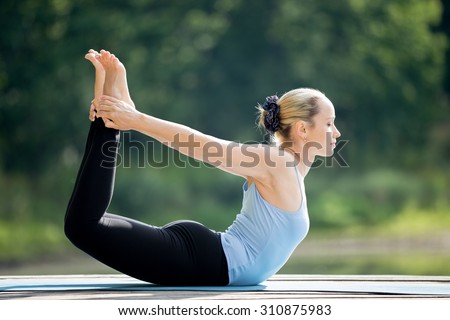 Beautiful sporty fit blond young woman wearing blue tank top sportswear working out outdoors on summer day, doing basic posture Dhanurasana, Bow Pose, full length