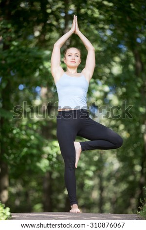 Beautiful fit young woman in blue tank top and black sporty leggings working out outdoors in park on summer day, standing in Tree posture (Vrkshasana), exercise for spine, full length