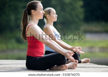 Two beautiful sporty fit young women sitting in Easy (Pleasant Posture), Sukhasana, meditating with closed eyes, breathing, working out outdoors in summer, wearing red and blue tank tops sportswear