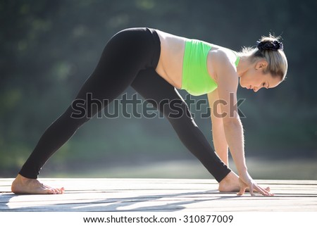 Beautiful sporty fit blond young woman in green sportswear working out outdoors, standing in Half Pyramid Pose, Intense Side Stretch Posture, Ardha Parsvottanasana, full length