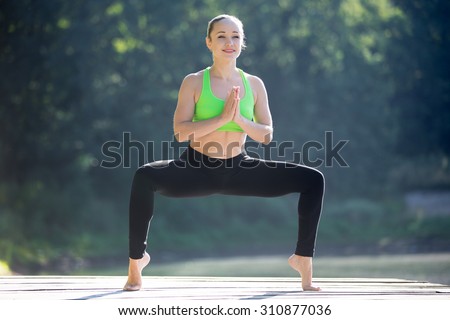 Beautiful sporty fit smiling blond young woman working out outdoors on wooden deck, standing in utkata konasana, rudrasana posture, full length