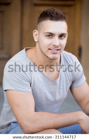 Portrait of hot guy looking in camera with a smile, wearing casual clothing, sitting in front of the wooden door on the street