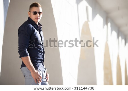 Portrait of young handsome man wearing blue shirt and sun glasses standing on the street beside white stucco wall with arched passageways, posing, copy space