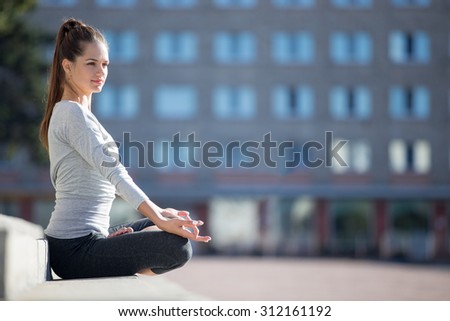 Yoga in the city: beautiful young fit woman meditating, breathing, sitting in Half Lotus Posture in front of building on the street on summer day, Ardha Padmasana, copy space
