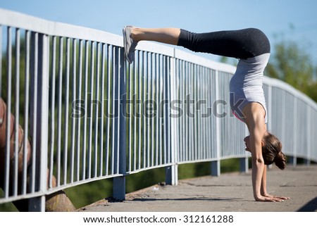 Yoga in the city: beautiful sporty young woman working out on the old bridge, leaning on railing in variation of Adho Mukha Vrkshasana with bent legs, Downward facing Tree Posture