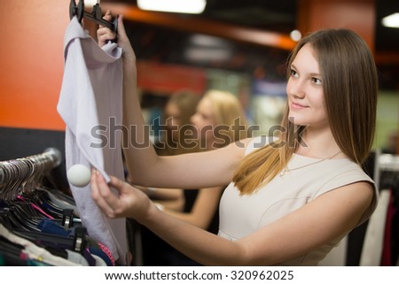 Portrait of smiling young beautiful woman shopping, standing in the mall, looking through clothes, holding hanger with blouse, choosing new outfit