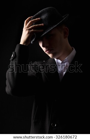 Handsome unrecognizable elegant mafioso man in coat touching his hat as in greeting, scarcely visible in the darkness