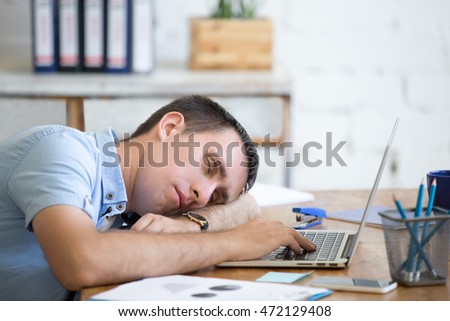 Portrait of young man lying on the table in front of laptop, sleepy, tired, overworked or lazy to work. Attractive business man napping in his home office relaxing after work on laptop computer