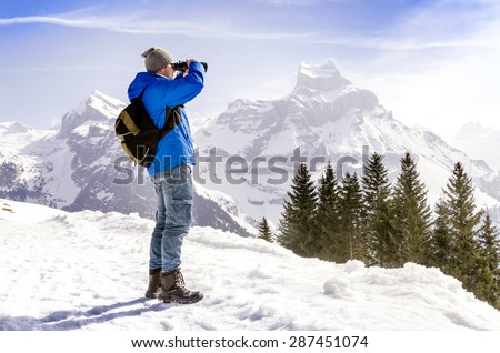 Man with backpack is standing on a mountain and looking through binoculars.